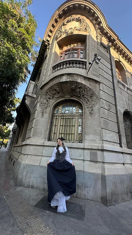 Persona de pie frente a una fachada antigua ornamentada, usando blusa blanca de mangas largas, corpiño negro y falda negra amplia con capa inferior blanca visible.