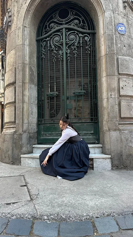 Persona sentada frente a una gran puerta de fierro verde, con vestido negro de falda voluminosa, corpiño oscuro y blusa blanca de mangas largas.