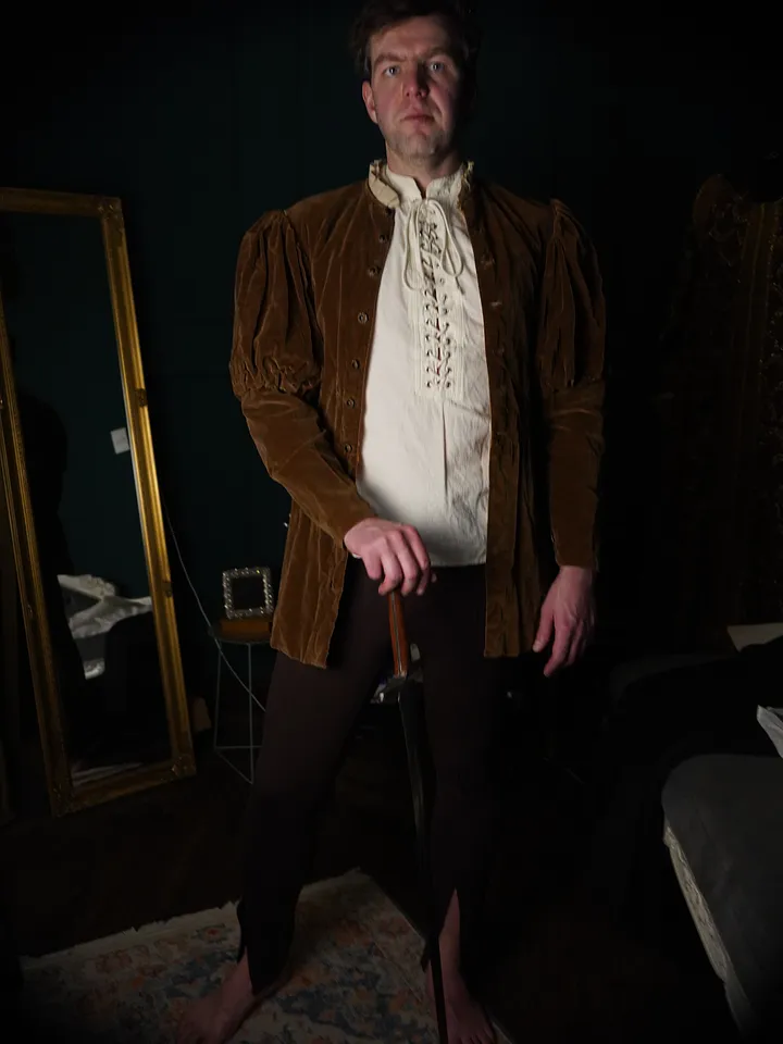 Hombre de frente con chaqueta de terciopelo ocre, hombros fruncidos, camisa marfil entrelazada y calzas oscuras.