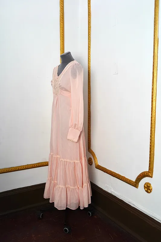Vista lateral del vestido rosado, mostrando la tela semitransparente, la cintura alta, la manga larga con puño ancho y los vuelos de la falda.