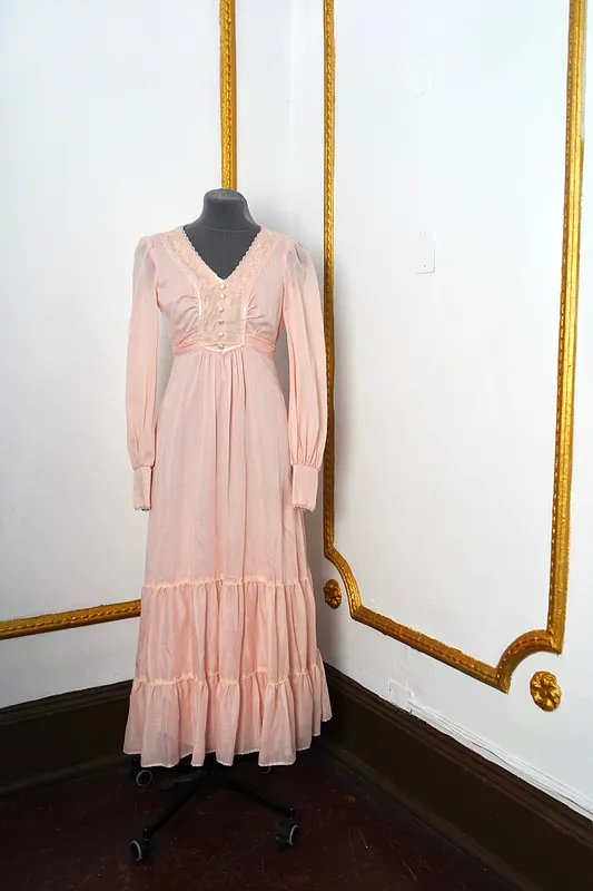 Vista frontal de vestido largo rosado en maniquí, con escote en V, encaje claro, botones forrados, mangas largas abullonadas y falda con vuelos.
