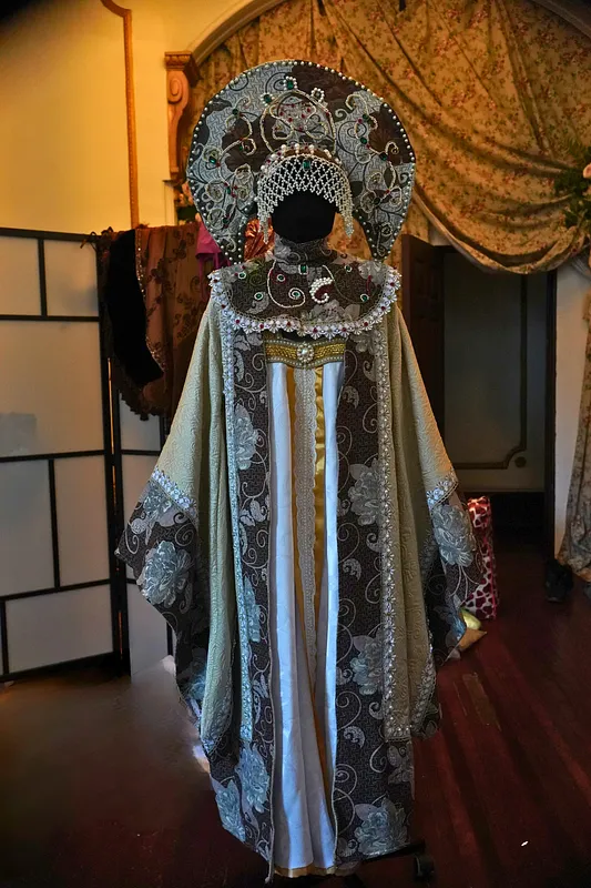 Vista frontal del traje en maniquí, con vestido largo brocado, mangas amplias marfil y gran tocado semicircular decorado con perlas y pedrería.