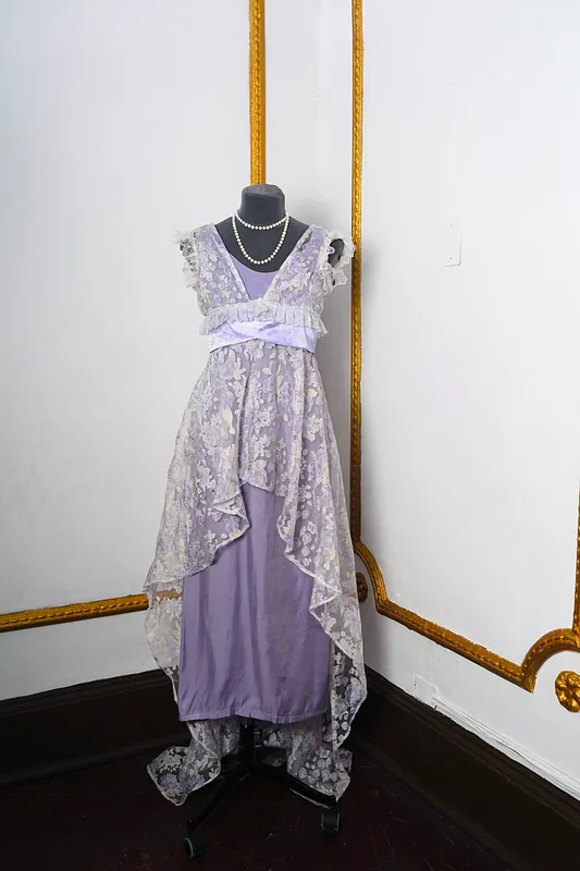 Vista frontal del vestido lavanda en maniquí, con escote en V, encaje floral translúcido, mangas con volantes, franja satinada y collar de perlas.