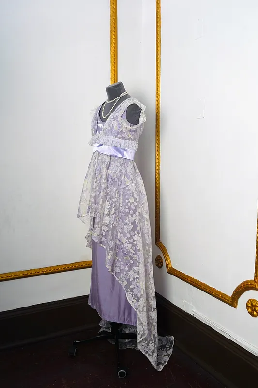 Vista lateral del vestido, mostrando la caída asimétrica de la capa de encaje floral sobre la falda lisa lavanda y la cintura satinada.