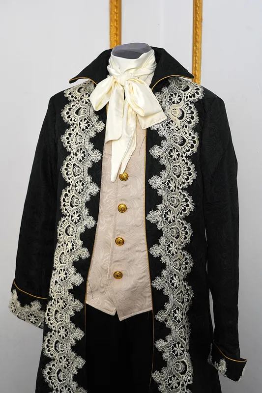 Plano medio del torso con solapas negras abiertas, lazo marfil al cuello, chaleco claro con botones dorados y encaje ornamental sobre la chaqueta.