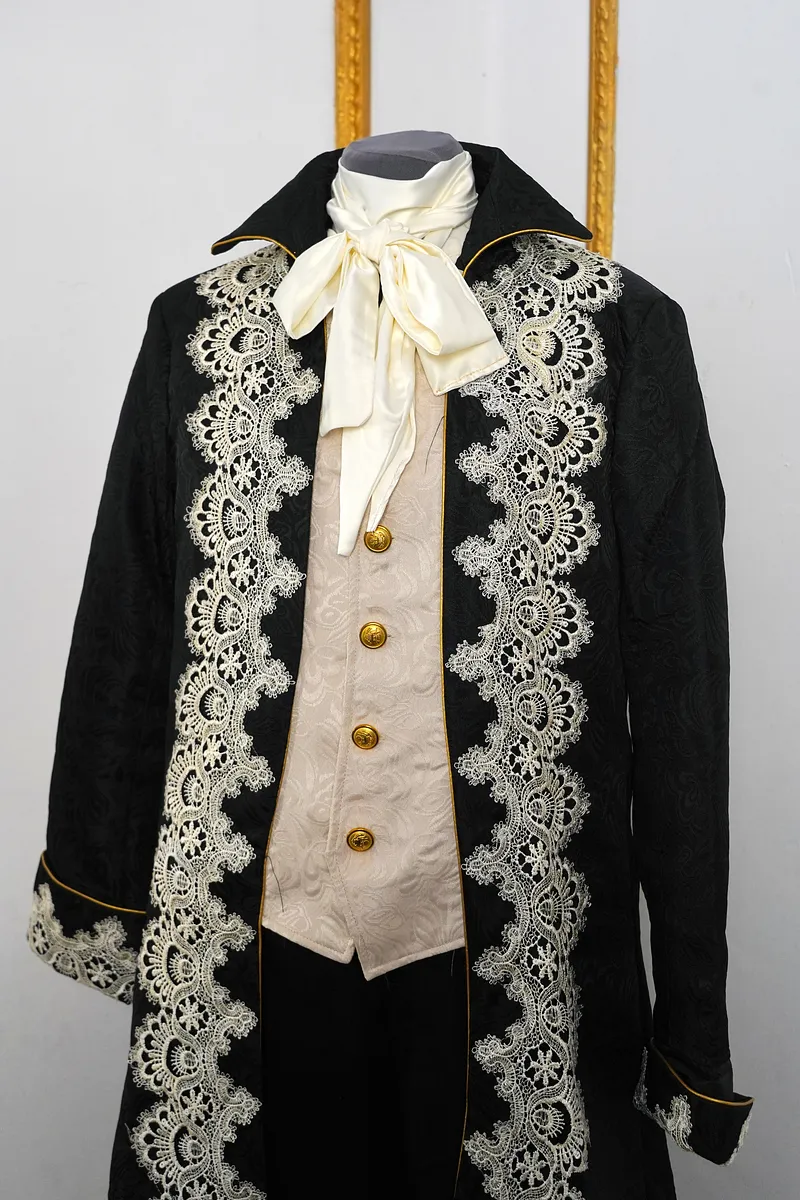 Plano medio del torso con solapas negras abiertas, lazo marfil al cuello, chaleco claro con botones dorados y encaje ornamental sobre la chaqueta.