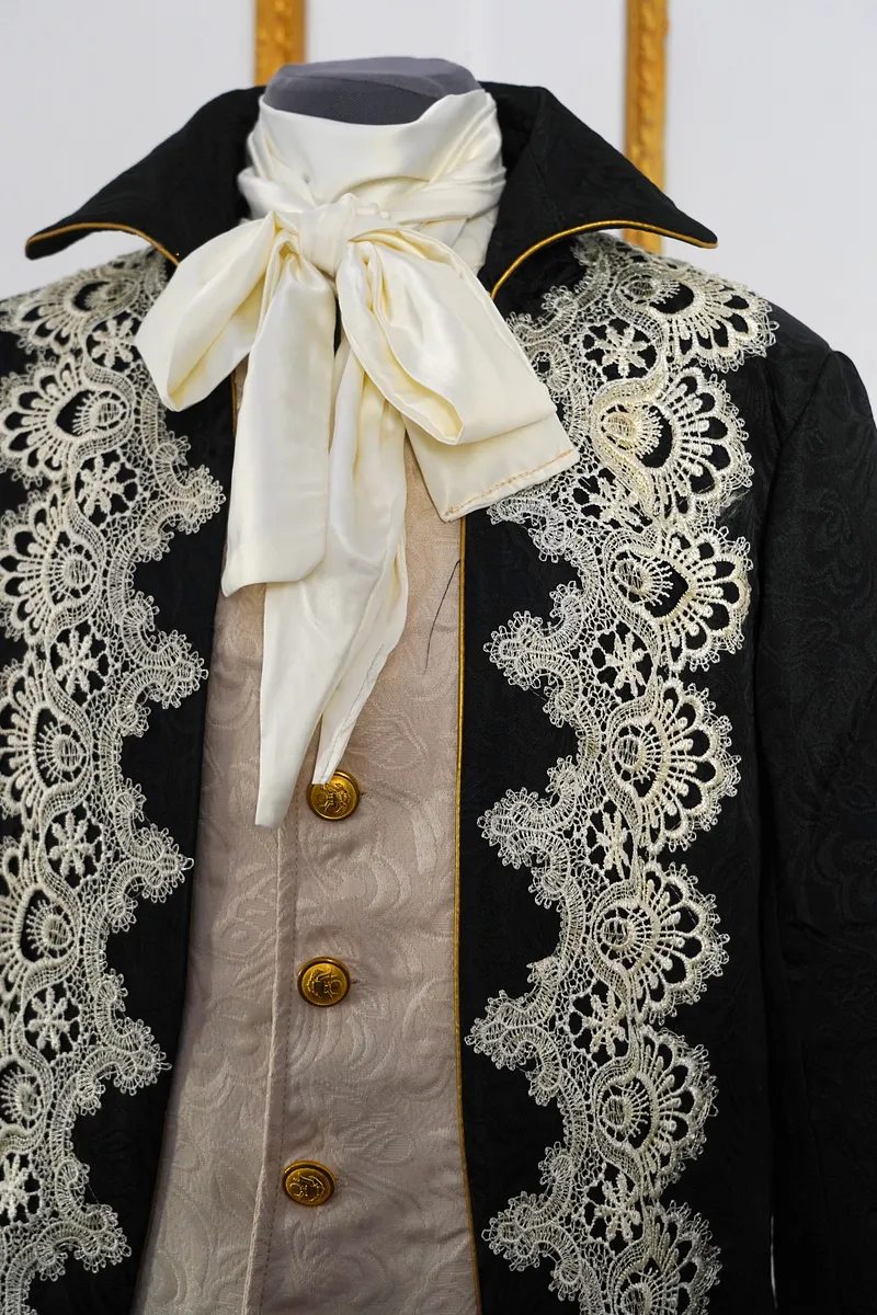 Detalle cercano del cuello y pecho, mostrando el lazo marfil satinado, el vivo dorado de la solapa, el chaleco brocado y el encaje aplicado.