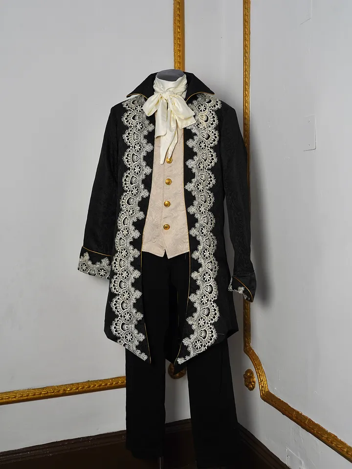 Vista frontal completa del conjunto en maniquí, con chaqueta negra larga, encaje marfil en los delanteros, chaleco claro, lazo al cuello y pantalón negro.