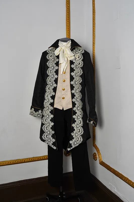 Vista frontal completa del conjunto en maniquí, con chaqueta negra larga, encaje marfil en los delanteros, chaleco claro, lazo al cuello y pantalón negro.