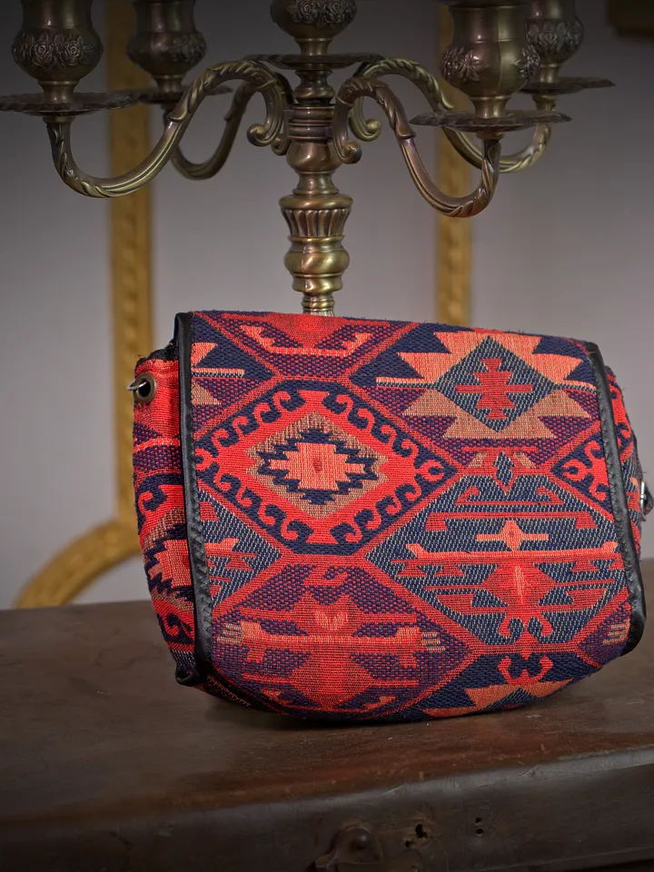 Cartera rectangular pequeña con tejido geométrico rojo, azul y negro, apoyada sobre un mueble de madera oscura.