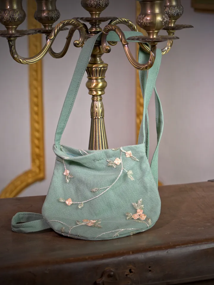 Bolso pequeño verde agua con asa corta, aplicaciones florales claras y detalles bordados, apoyado sobre una superficie de madera.