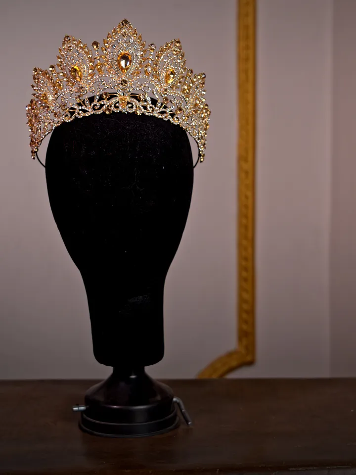 Corona dorada abierta sobre soporte negro, con puntas ornamentadas, filigrana y numerosas piedras transparentes de distintos tamaños.