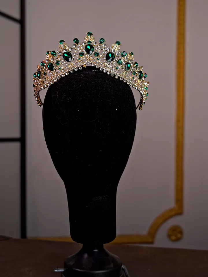 Tiara plateada sobre soporte negro, decorada con piedras verdes ovaladas, cristales transparentes y pequeñas puntas ornamentales.
