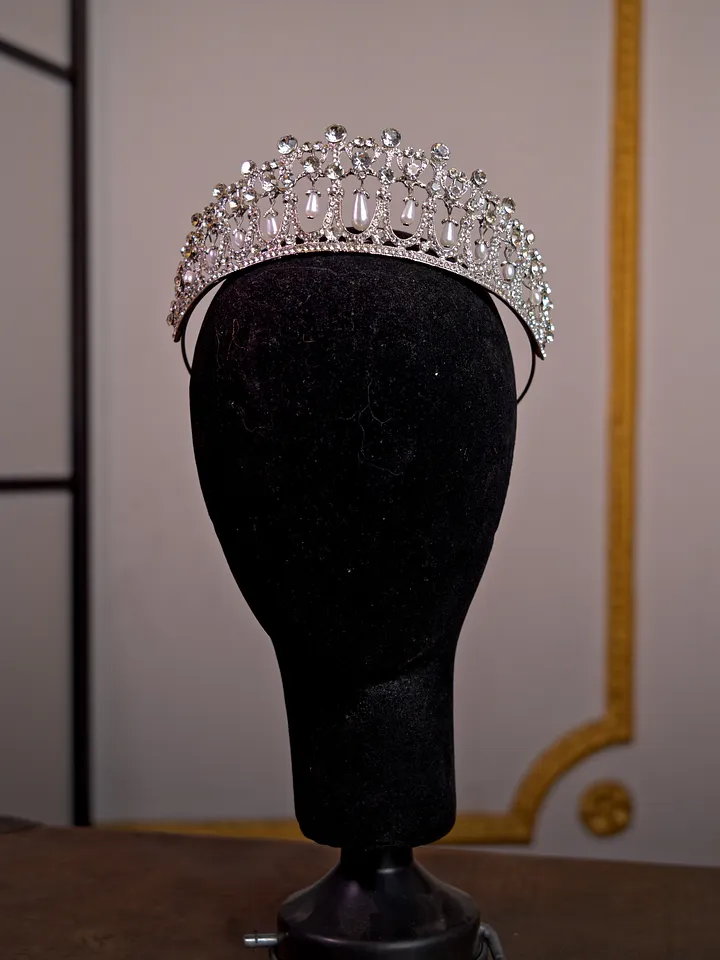Tiara plateada de cristales transparentes sobre cabeza de maniquí, con arcos simétricos y una hilera superior de pequeñas piedras redondas.