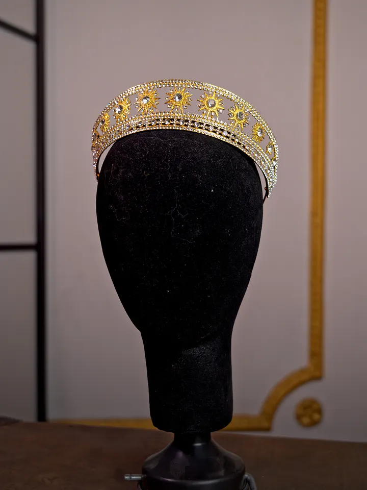 Corona dorada de perfil bajo sobre soporte negro, decorada con filigrana, piedras transparentes y pequeños detalles de tono violeta.