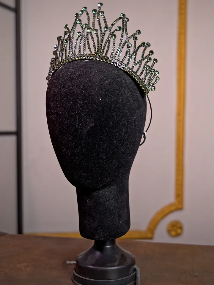 Tiara plateada alta con estructura de puntas curvas y pequeñas piedras verdes, presentada sobre una cabeza de maniquí negra.