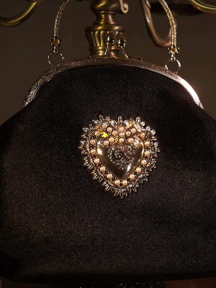 Pequeño bolso negro de tela con cierre metálico, asa corta y broche central en forma de corazón rodeado de pedrería.