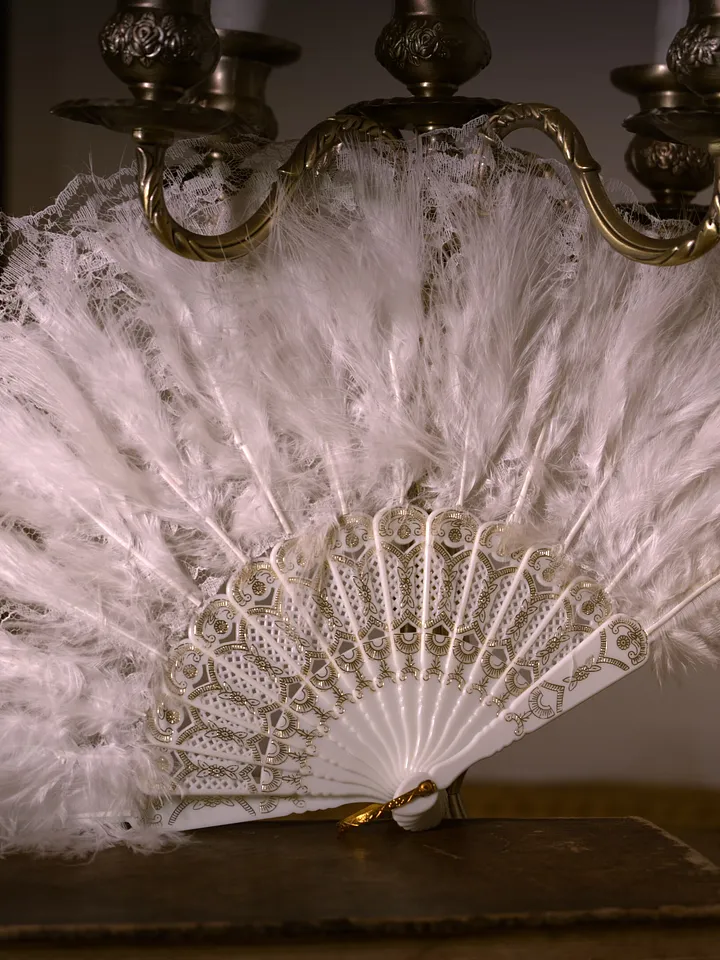 Abanico abierto de plumas blancas, con varillas claras decoradas y un borde superior de encaje festoneado.