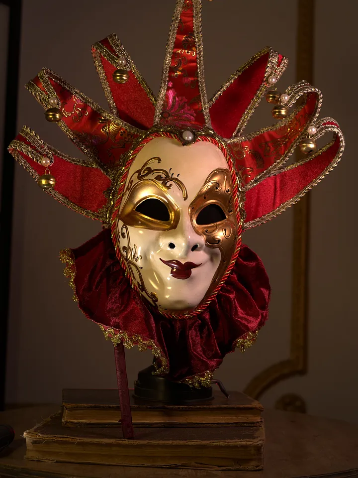 Máscara de arlequín sobre soporte, con rostro dorado y blanco, antifaz oscuro, cuello rojo y puntas rojas ribeteadas en dorado.