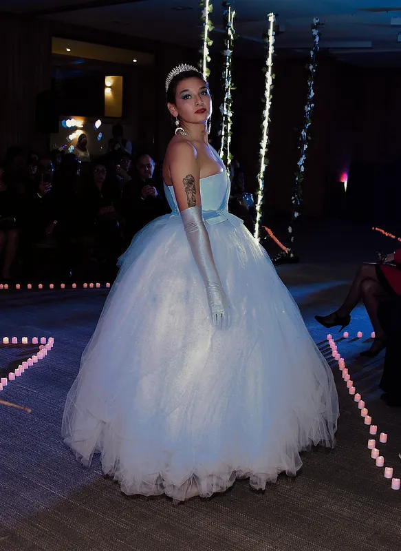 Modelo en pasarela interior usando un vestido celeste claro con corpiño strapless, cintura marcada y falda muy amplia de tul; lo combina con guantes largos y tiara.