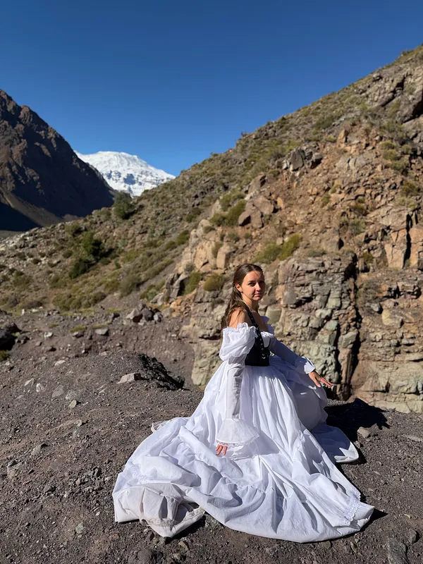 Mujer sentada sobre terreno rocoso con vestido blanco y corpiño negro, mientras la falda y la cola se extienden alrededor.