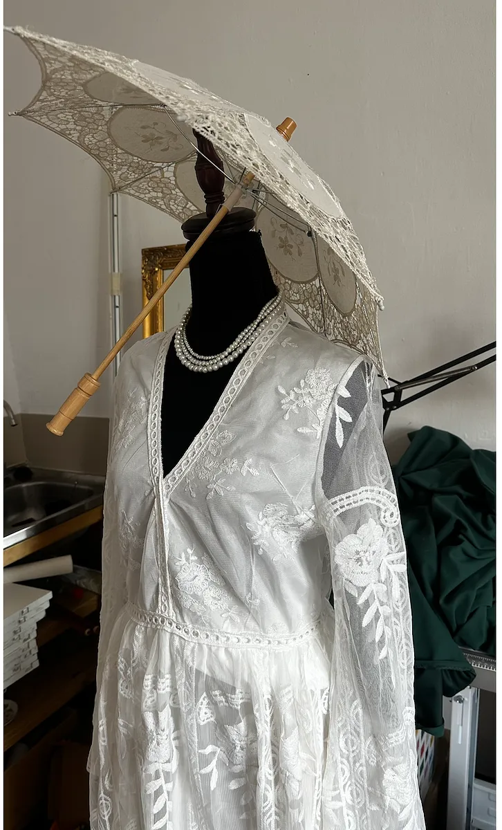 Acercamiento al cuerpo del vestido blanco, acompañado por collar de perlas y una sombrilla de encaje sostenida sobre el hombro.
