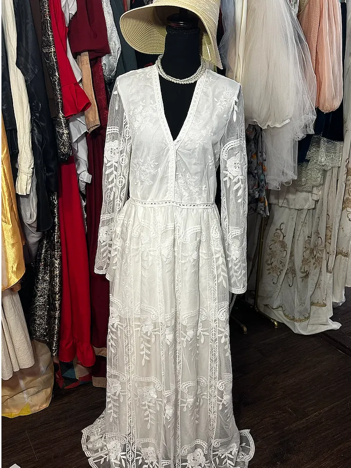 Vista frontal de vestido blanco largo de encaje, con escote en V, mangas transparentes acampanadas y sombrero claro.