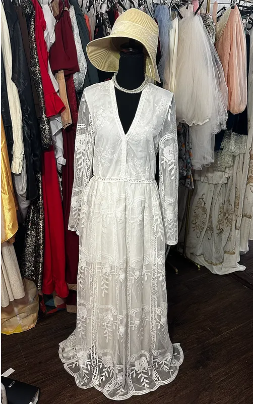 Vista frontal de vestido blanco largo de encaje, con escote en V, mangas transparentes acampanadas y sombrero claro.
