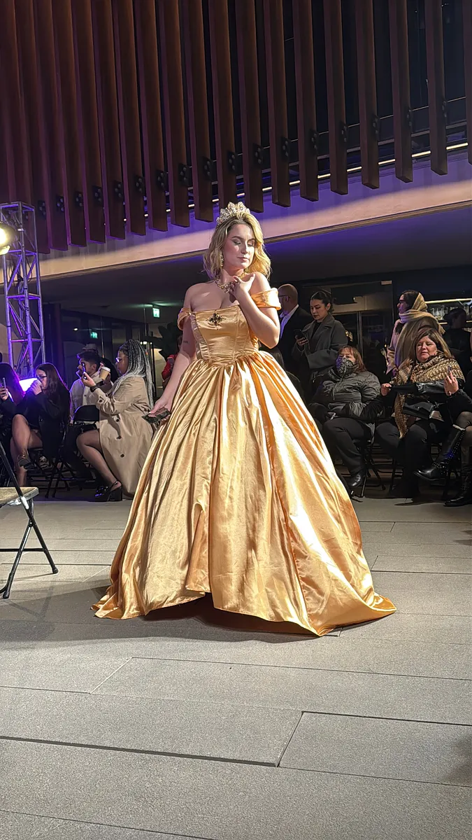 Mujer camina de frente por una pasarela con vestido dorado brillante, mangas caídas y falda amplia hasta el suelo.