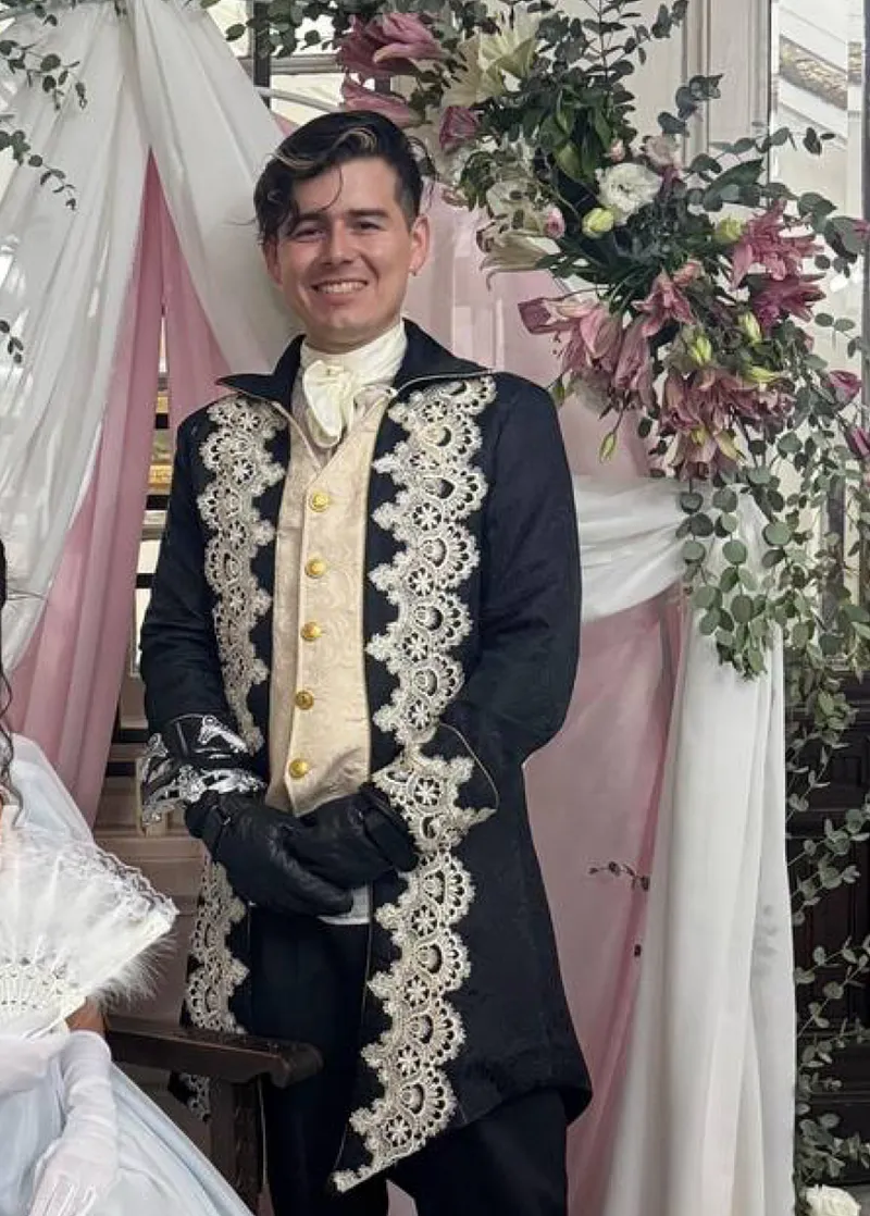 Persona usando el conjunto en un montaje decorado con flores y telas claras, con chaqueta negra de encaje, chaleco claro, guantes negros y lazo al cuello.