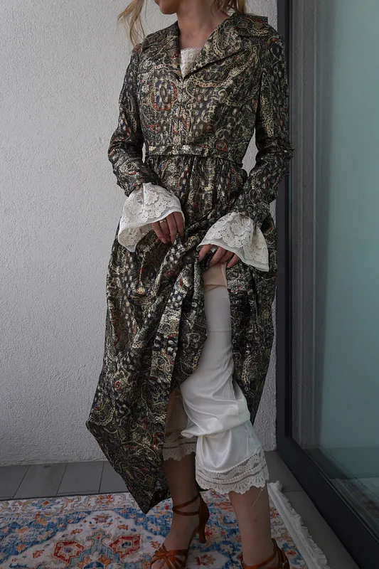 Vestido largo oscuro con brillo y estampado paisley, cuello con solapa, cintura marcada y mangas largas con puños amplios de encaje blanco.