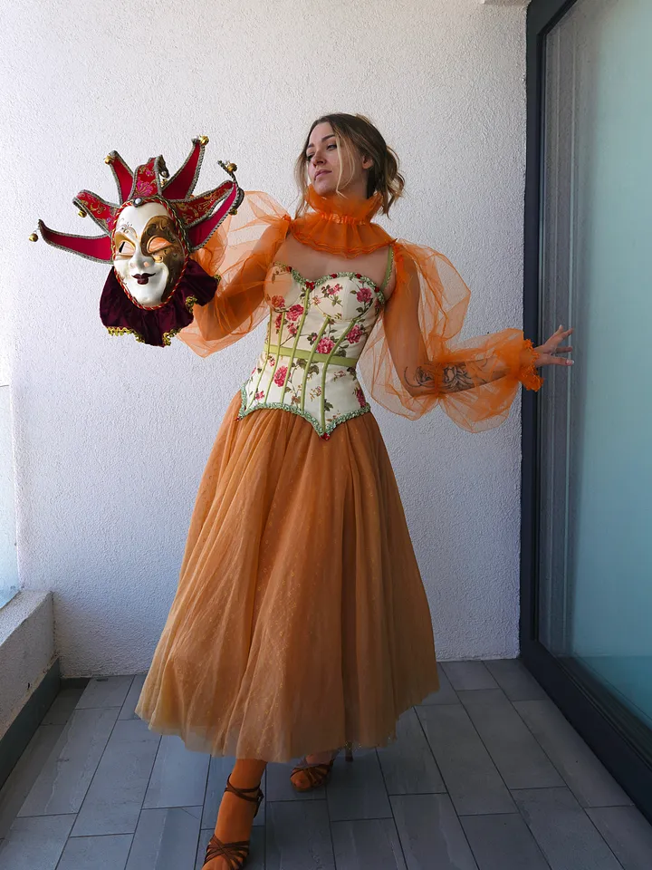 Mujer con vestido naranja de falda amplia de tul, mangas translúcidas abullonadas, cuello alto con volante y corpiño crema floral con ribetes verdes; sostiene una máscara veneciana decorativa.