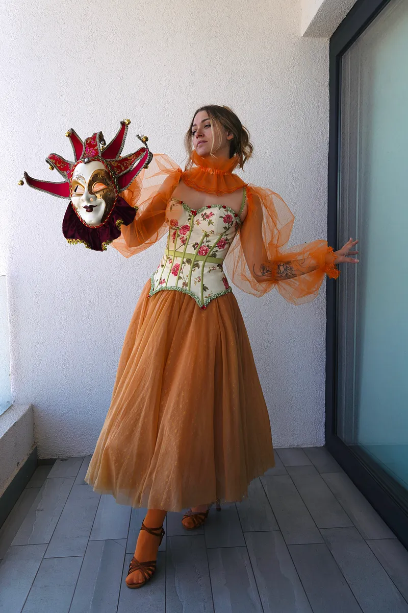 Mujer con vestido naranja de falda amplia de tul, mangas translúcidas abullonadas, cuello alto con volante y corpiño crema floral con ribetes verdes; sostiene una máscara veneciana decorativa.
