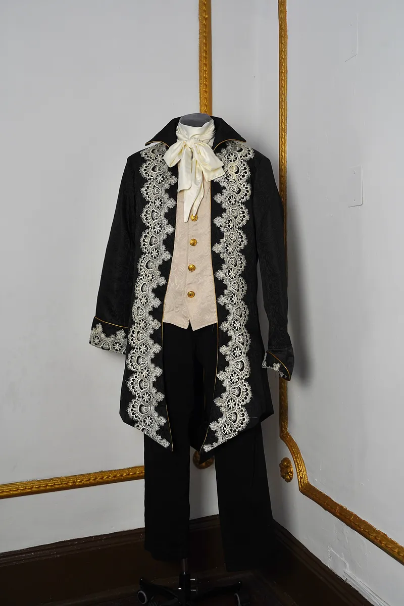 Vista frontal completa del conjunto en maniquí, con chaqueta negra larga, encaje marfil en los delanteros, chaleco claro, lazo al cuello y pantalón negro.