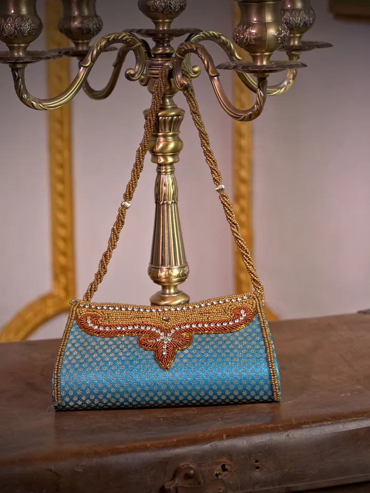 Cartera de fiesta turquesa con superficie de cuentas, detalle frontal anaranjado y dorado, y cadena metálica larga.
