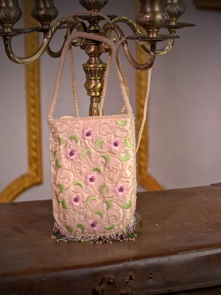 Bolso rectangular rosado con correa delgada, superficie bordada con pequeñas flores y aplicaciones brillantes, colgado frente a un muro claro.