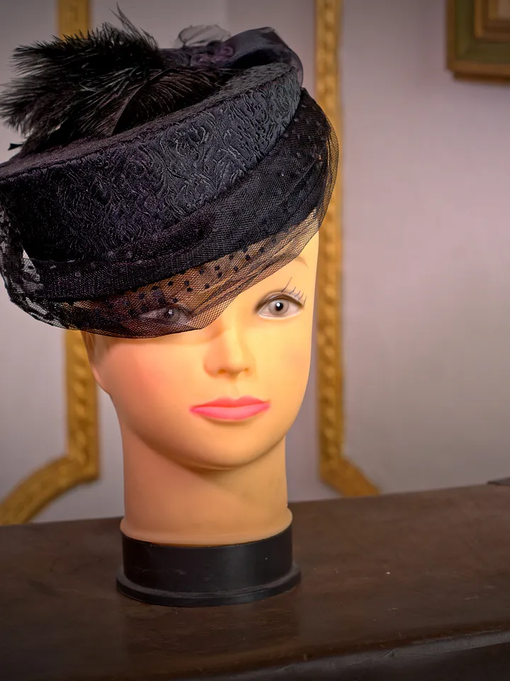 Sombrero negro pequeño sobre cabeza de maniquí, con copa baja, ala inclinada, cintas superpuestas y un adorno de plumas.