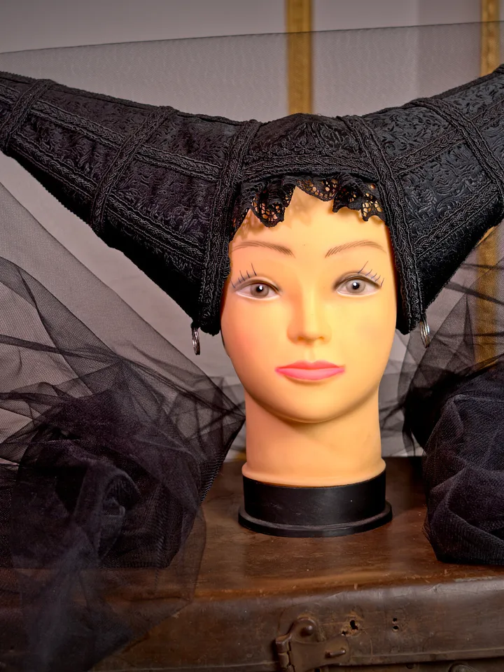 Tocado negro de silueta angular con dos puntas laterales, textura bordada y amplio velo de tul desplegado alrededor del maniquí.
