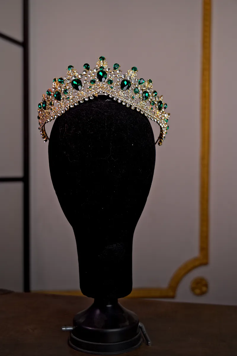 Tiara plateada sobre soporte negro, decorada con piedras verdes ovaladas, cristales transparentes y pequeñas puntas ornamentales.