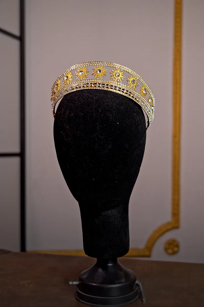 Corona dorada de perfil bajo sobre soporte negro, decorada con filigrana, piedras transparentes y pequeños detalles de tono violeta.