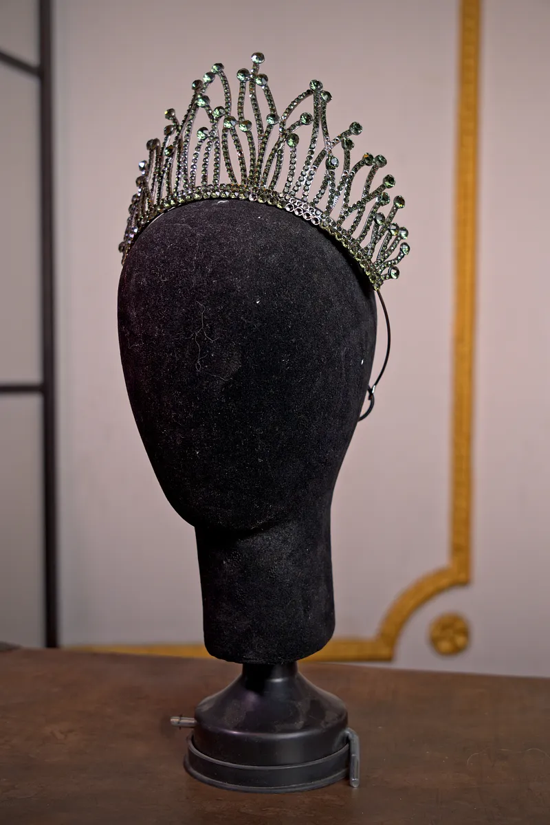 Tiara plateada alta con estructura de puntas curvas y pequeñas piedras verdes, presentada sobre una cabeza de maniquí negra.