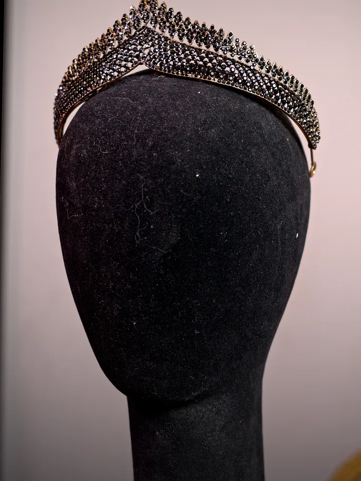 Tiara alta de metal oscuro con diseño calado y numerosas puntas, cubierta por pequeñas piedras brillantes en varias hileras.