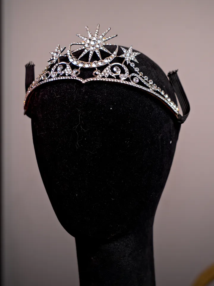 Tiara plateada con diseño asimétrico de estrellas y espirales, decorada con cristales transparentes sobre un soporte negro.