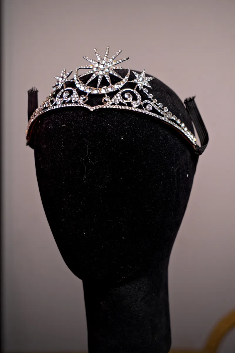 Tiara plateada con diseño asimétrico de estrellas y espirales, decorada con cristales transparentes sobre un soporte negro.