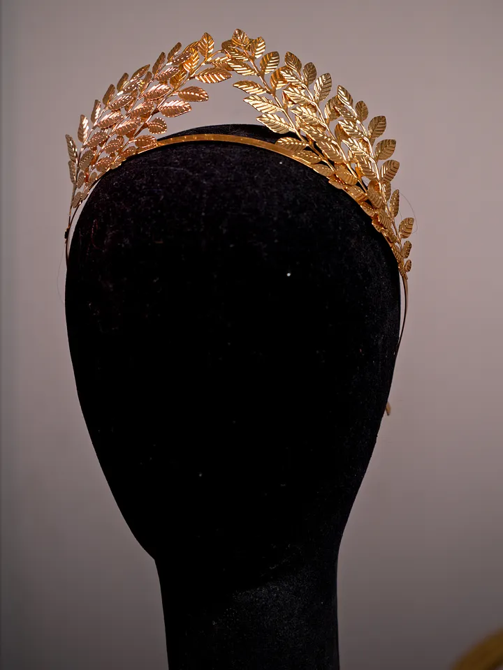 Tiara dorada formada por ramas de hojas de laurel superpuestas, presentada sobre una cabeza de maniquí negra.