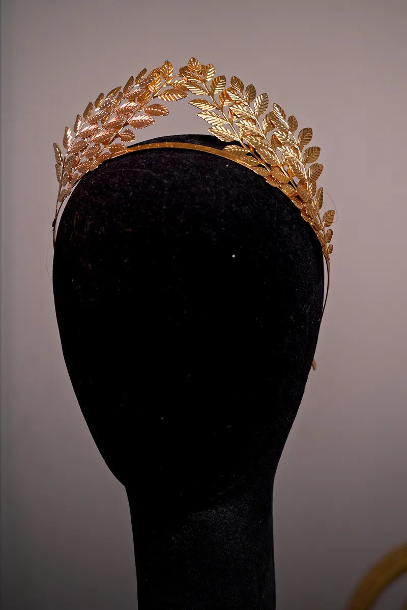 Tiara dorada formada por ramas de hojas de laurel superpuestas, presentada sobre una cabeza de maniquí negra.