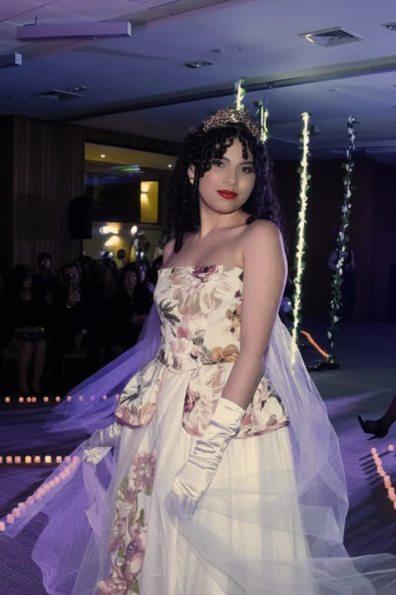 Modelo en pasarela interior con vestido strapless floral, sobrefalda peplum, falda blanca con capas de tul translúcido y guantes claros de estilismo.