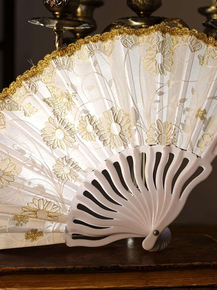 Abanico plegable blanco abierto, con borde dorado, varillas claras y motivos florales pálidos sobre la superficie.