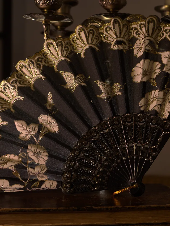 Abanico negro abierto con varillas oscuras y decoración floral dorada y crema concentrada a lo largo del borde superior.