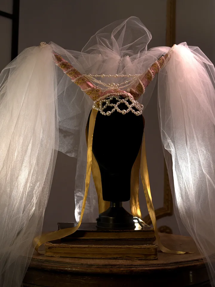 Tocado de fantasía sobre maniquí, con grandes capas blancas de tul y plumas extendidas, sujetas por cintas doradas y adorno central.